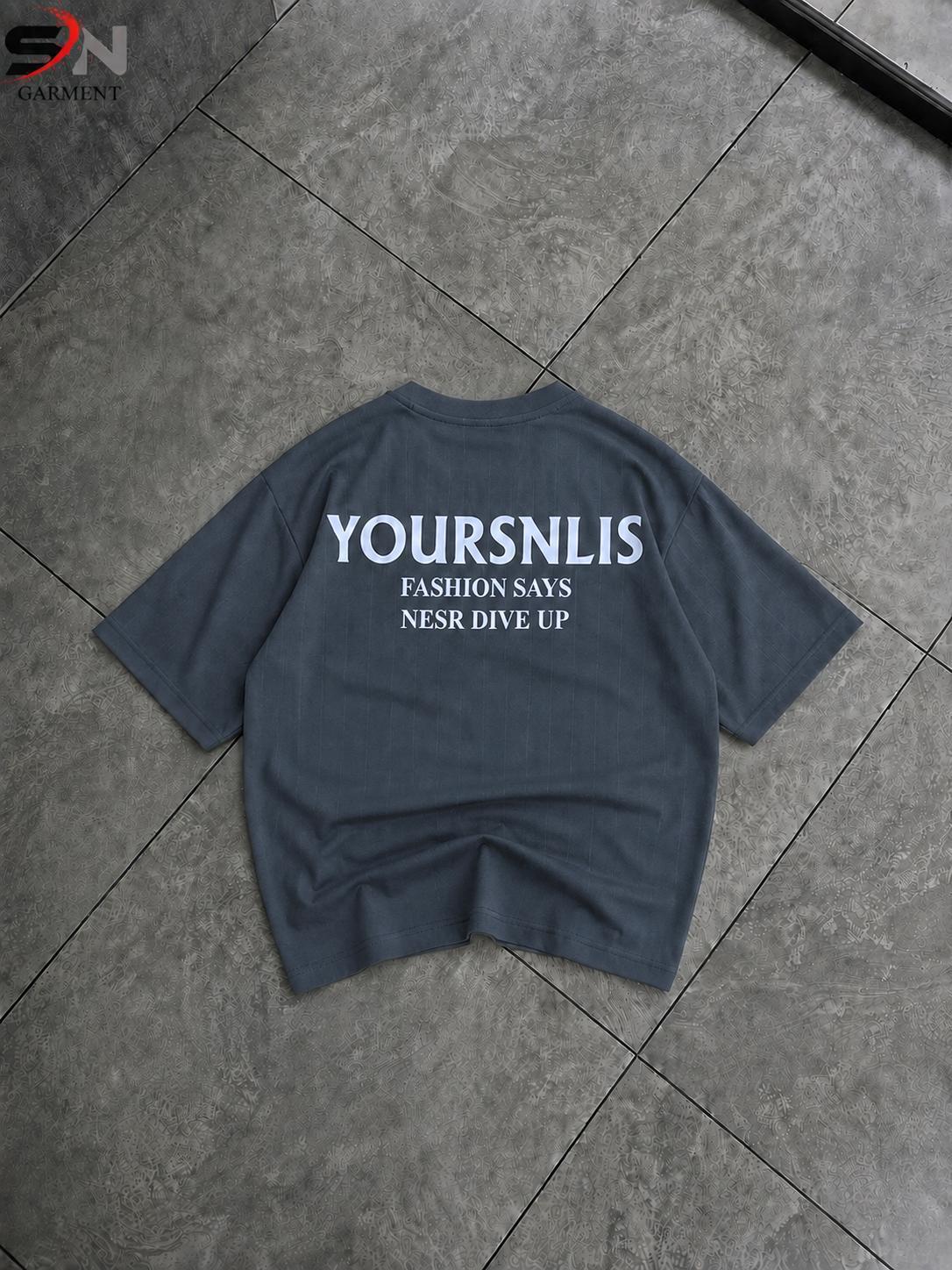A dark slate blue-gray t-shirt is laid flat on a gray tiled floor, showing the back side with white printed text that reads 'YOURSNLIS FASHION SAYS NESR DIVE UP'.