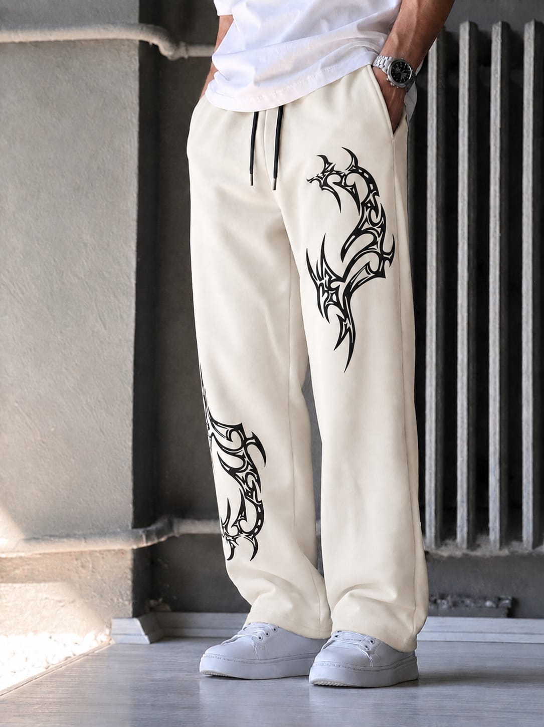 A person wearing off-white drawstring sweatpants with large black tribal-style graphic prints on both legs, paired with a white t-shirt and white sneakers, standing in an industrial setting.