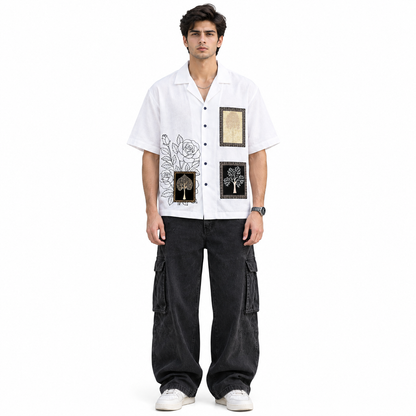A man modeling a white short-sleeve button-up shirt with black and gold graphic prints, paired with black cargo pants and white sneakers, against a plain white background.